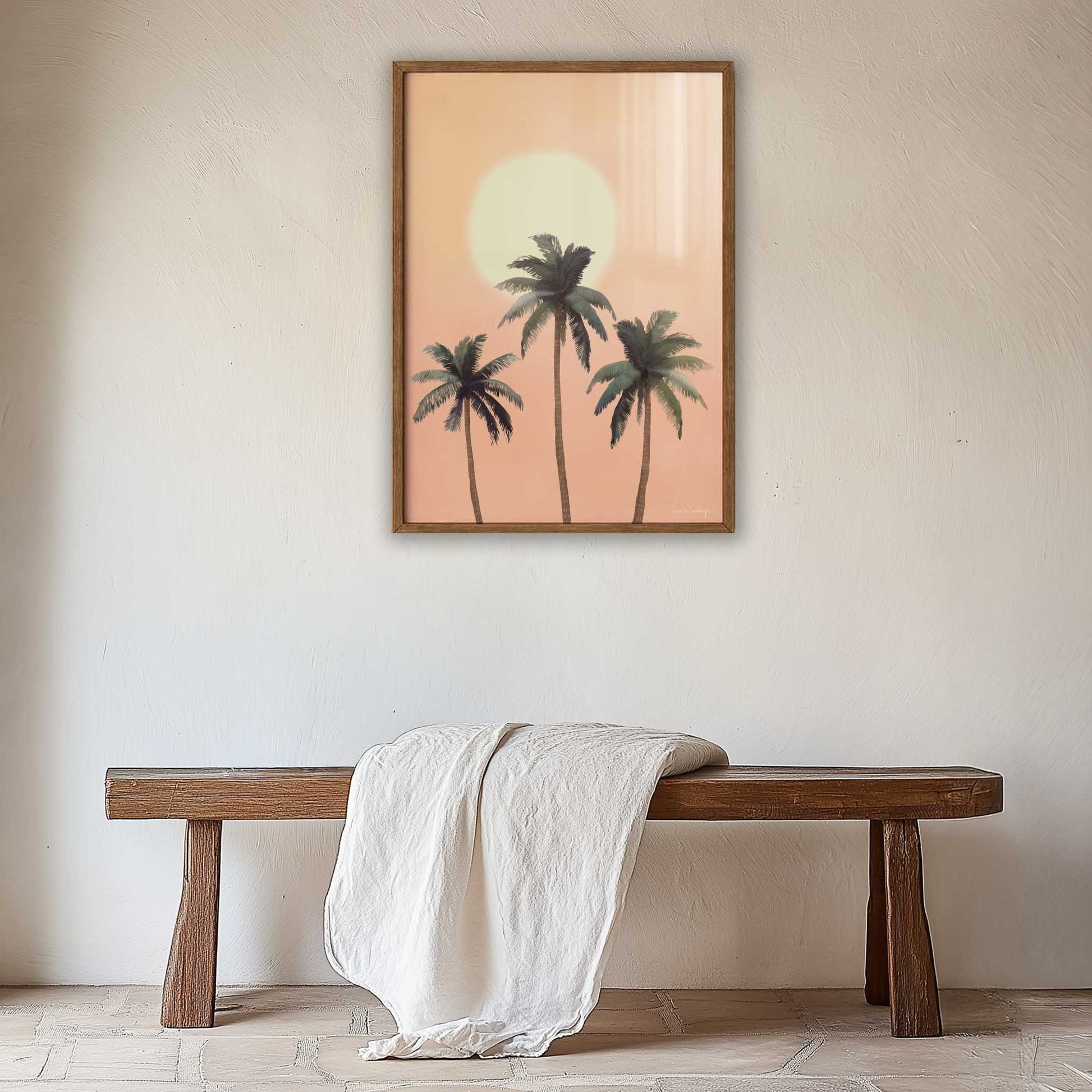 Wooden bench with a white towel draped over it, against a wall with a framed artwork of palm trees and a sunset.