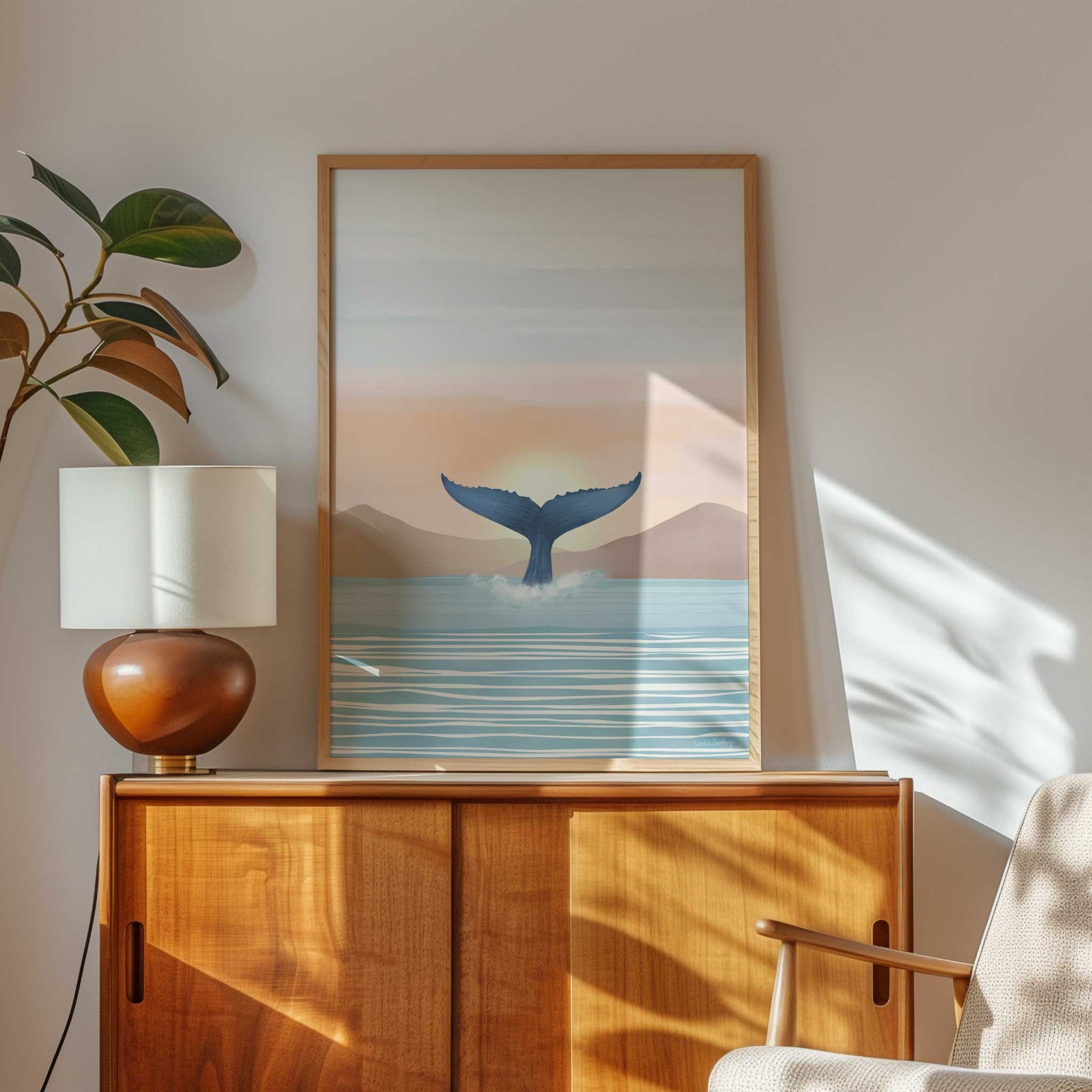 Framed artwork of a whale tail over water with a lamp and plant on a wooden sideboard.